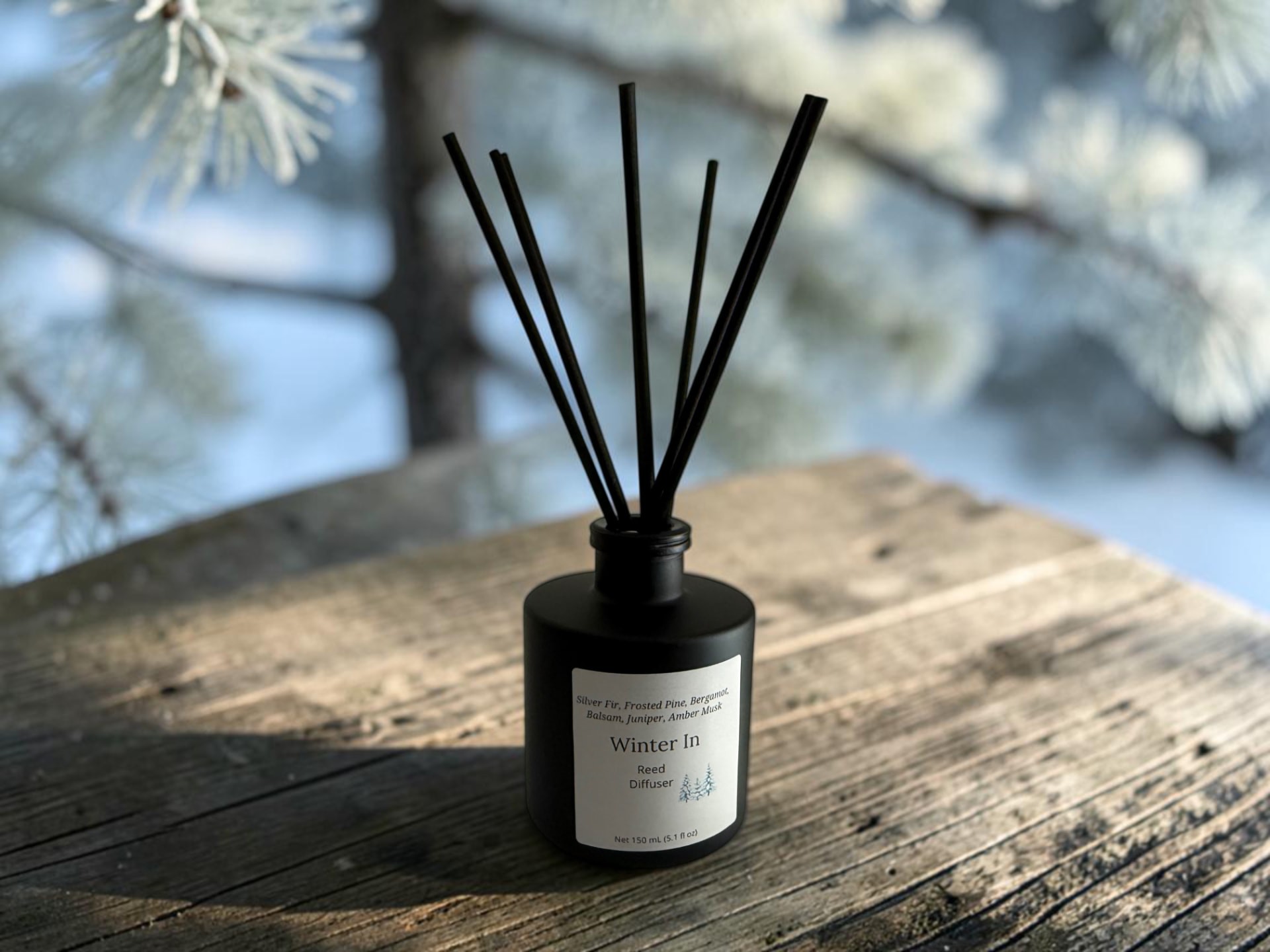 Winter In Reed Diffuser, a matte black bottle with black reeds set against a snowy evergreen forest