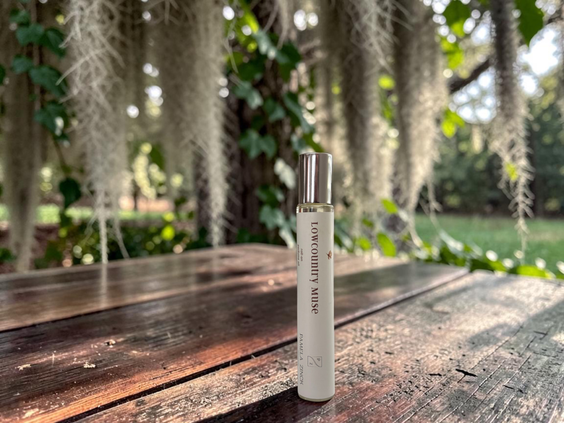 Lowcountry Muse Perfume Oil, a slim rollerball bottle standing on a weathered wooden table outdoors with Spanish moss and trees in the background