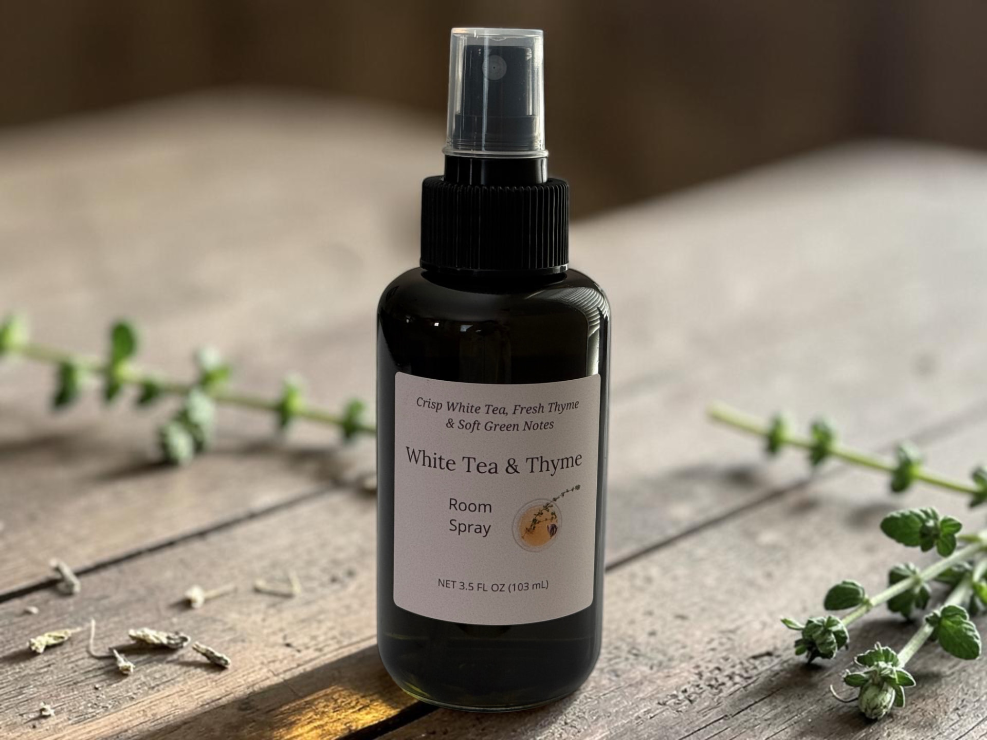 White Tea & Thyme Room Spray, a black bottle with a clear spray cap on weathered wooden planks beside fresh thyme sprigs and white tea leaves