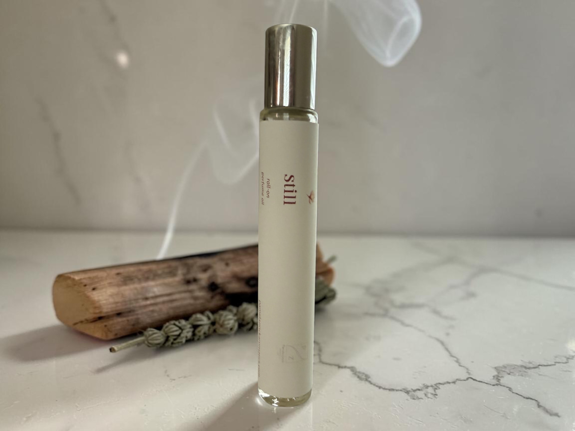 Still Perfume Oil, a slim rollerball bottle with a silver cap standing on a marble counter beside dried lavender and a wooden tool