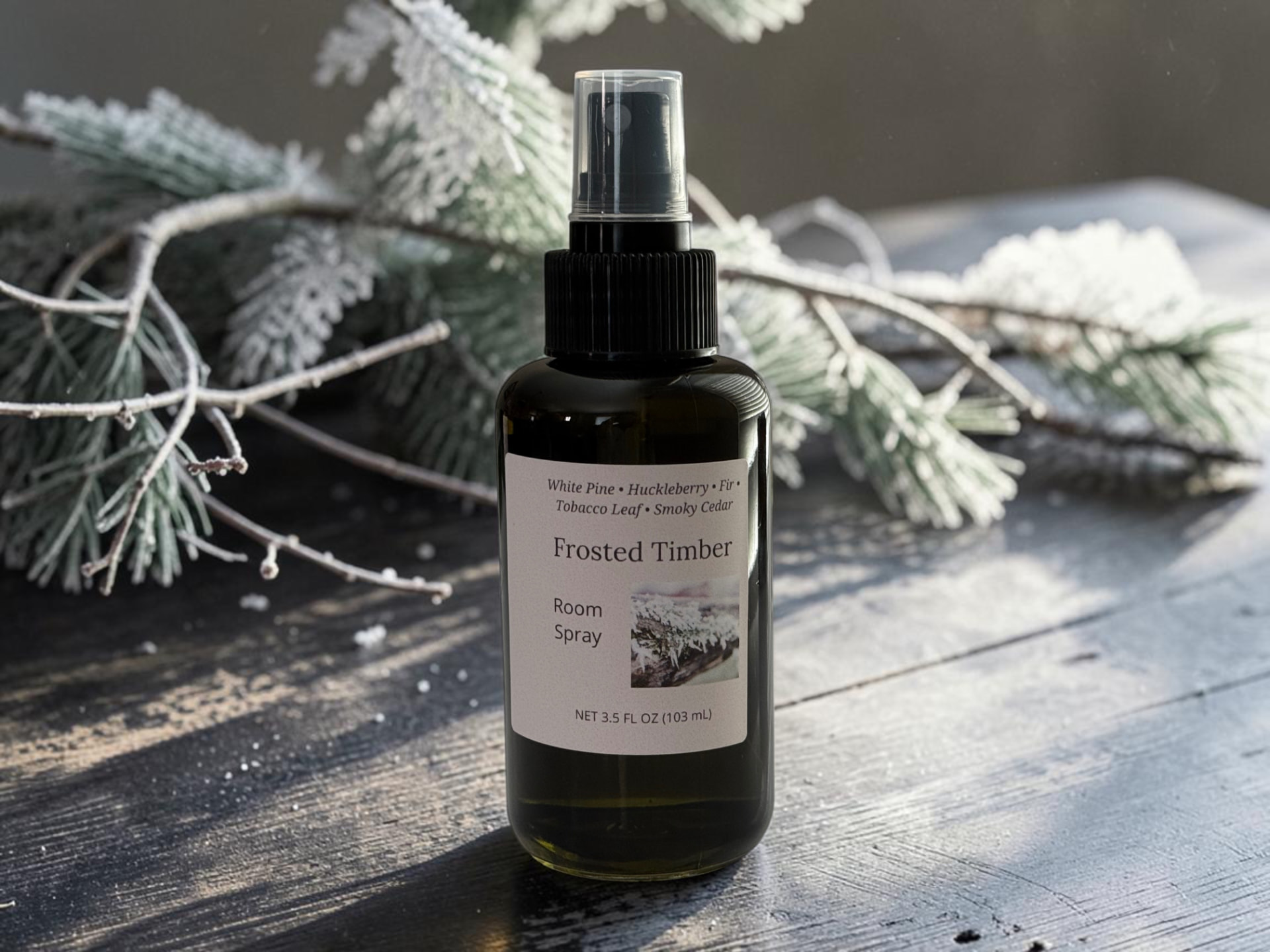 Frosted Timber Room Spray, a black bottle with a clear spray cap standing on weathered wooden planks with frosted evergreen branches in the background