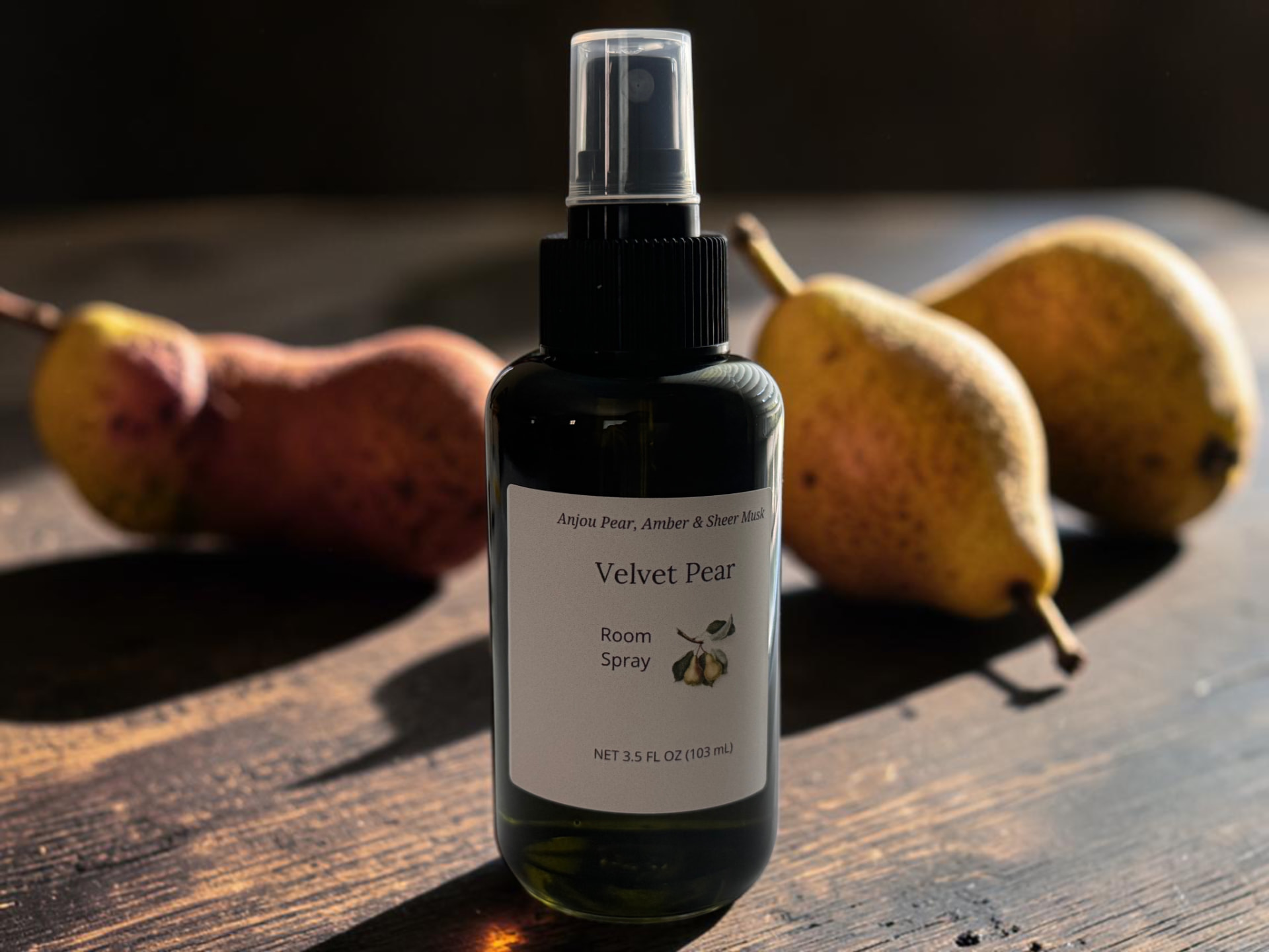Velvet Pear Room Spray, a black spray bottle with Anjou pear, amber, and sheer musk notes, set among ripe pears on a wooden surface