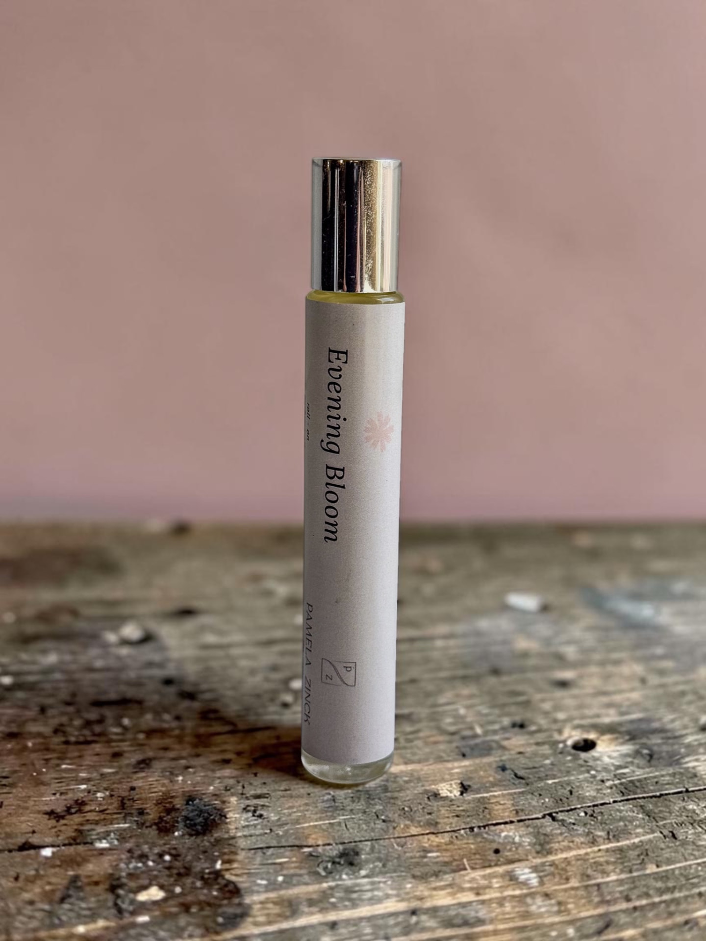 Evening Bloom Perfume Oil, a slim rollerball bottle with a gold cap standing on a weathered wooden table against a soft pink background