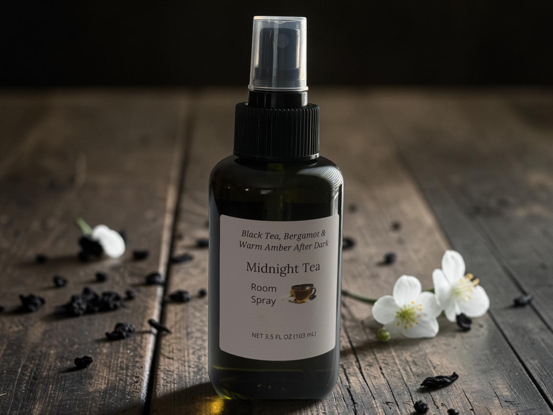 Midnight Tea Room Spray, a black bottle with a clear spray cap on weathered wooden planks beside white jasmine blossoms and loose black tea leaves